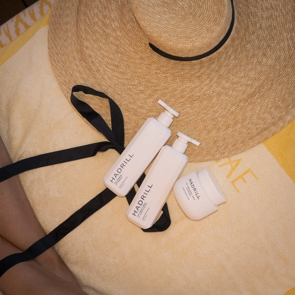 Hadrill Shampoo, Conditioner and Treatment Mask on a yellow beach towel beside a straw hat
